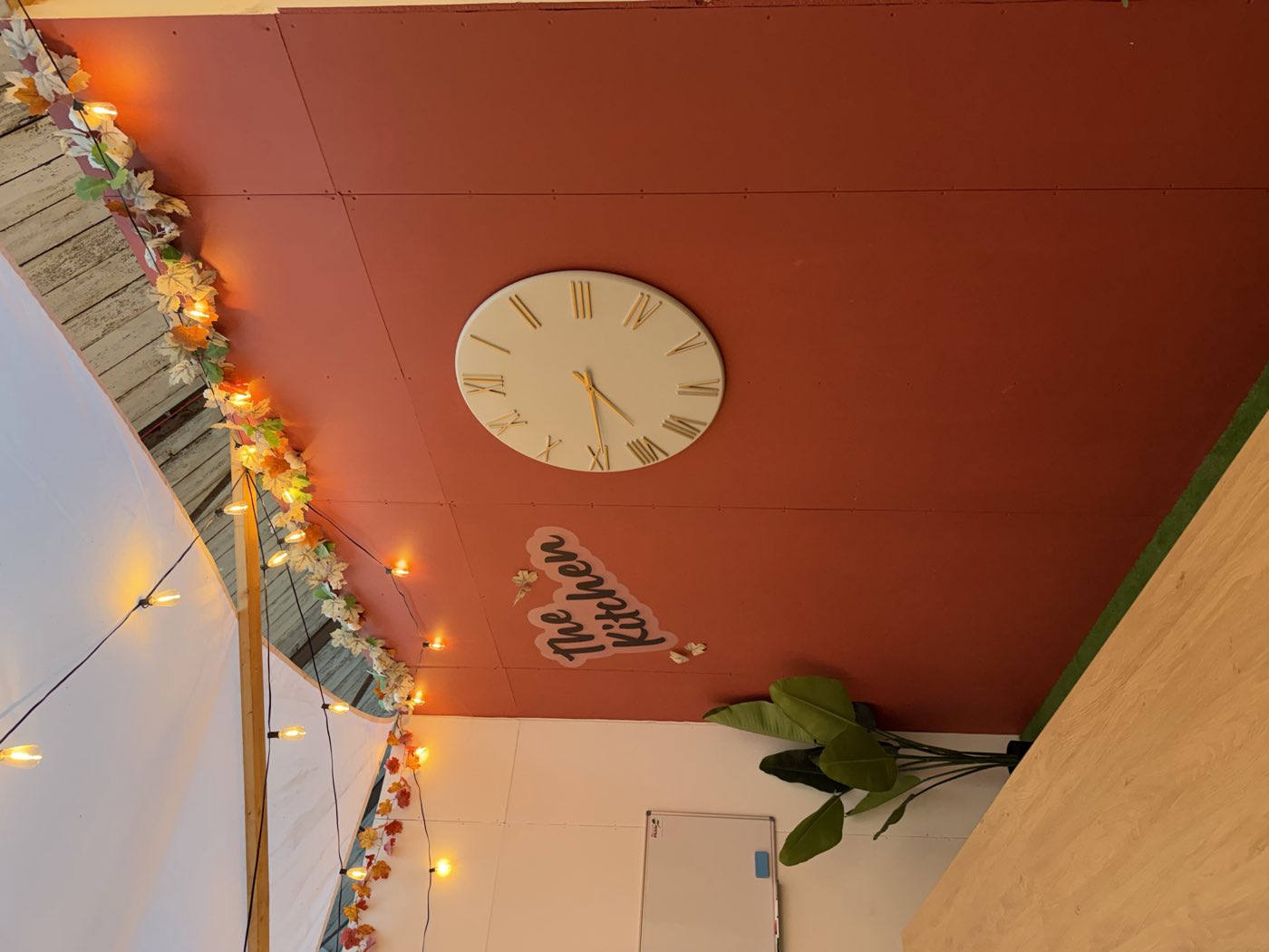 The Kitchen at The Court Yard: a terracotta wall with a painted sign and a large cream clock, under strings of warm bulbs and autumn garlands