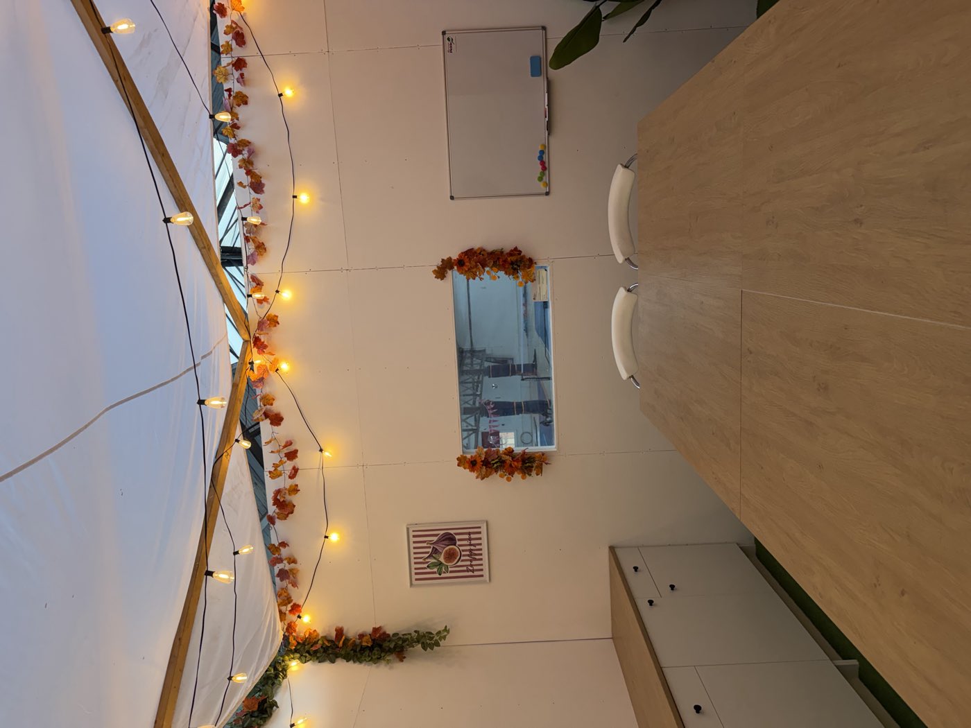 A long oak table set for a group under strings of warm bulbs, with a window through to the courts behind